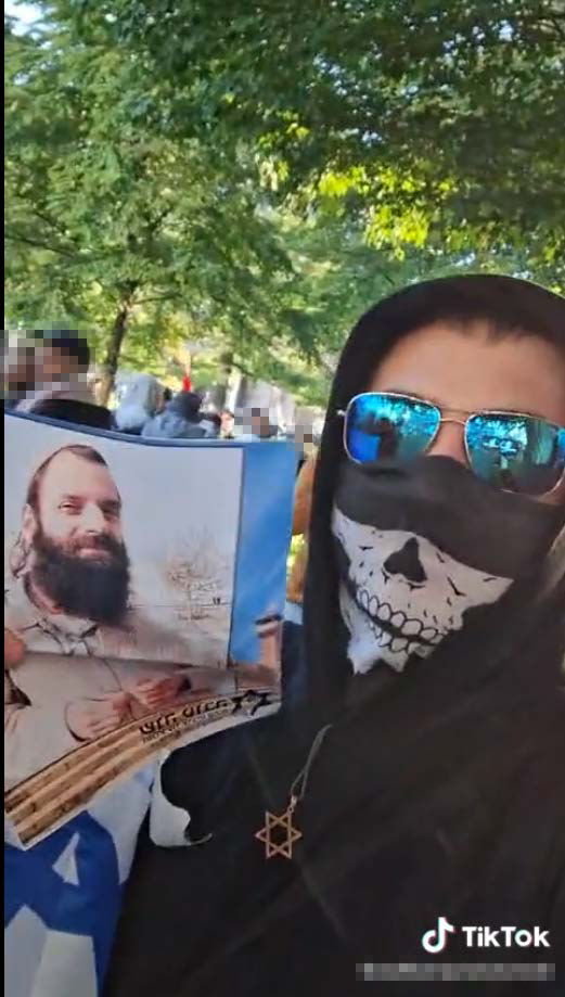 A boy or young man adorning a skull balaclava under aviator sunglasses and a Star of David necklace. stands in front of a crowd of people with blurred faces in daylight, with trees around. He holds up a picture of a man (Baruch Goldstein) smiling, aimed towards the camera. The TikTok watermark is present on the bottom-right.